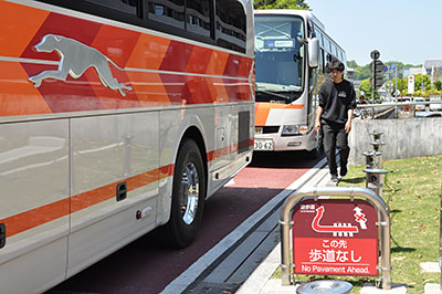 歩道が撤去された奈良市道で信号待ちのため停止した観光バスとその脇の路肩を歩く通行者。この路肩の部分に歩道があった。右手前は、歩道に代わる県文化会館広場通路への誘導看板＝2019年5月4日、奈良市登大路町