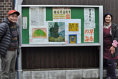 掲示板の横に立つ奥田さん（右）と絵画教室を開いている横堀さん＝同