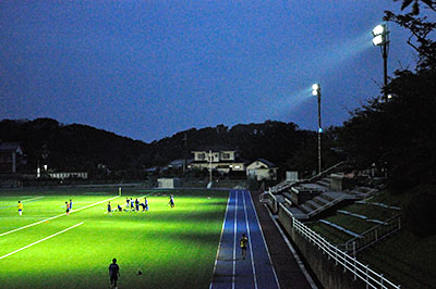夜間照明（右）が点灯された日没直後の生駒北スポーツセンターのグラウンド。奥は照明の光を受ける住宅